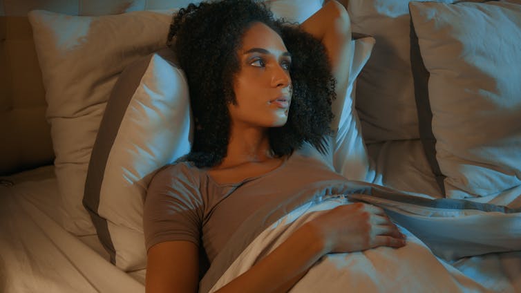 Jovem adulta deitada na cama, bem acordada, olhando pela janela em direção a uma luz brilhante.